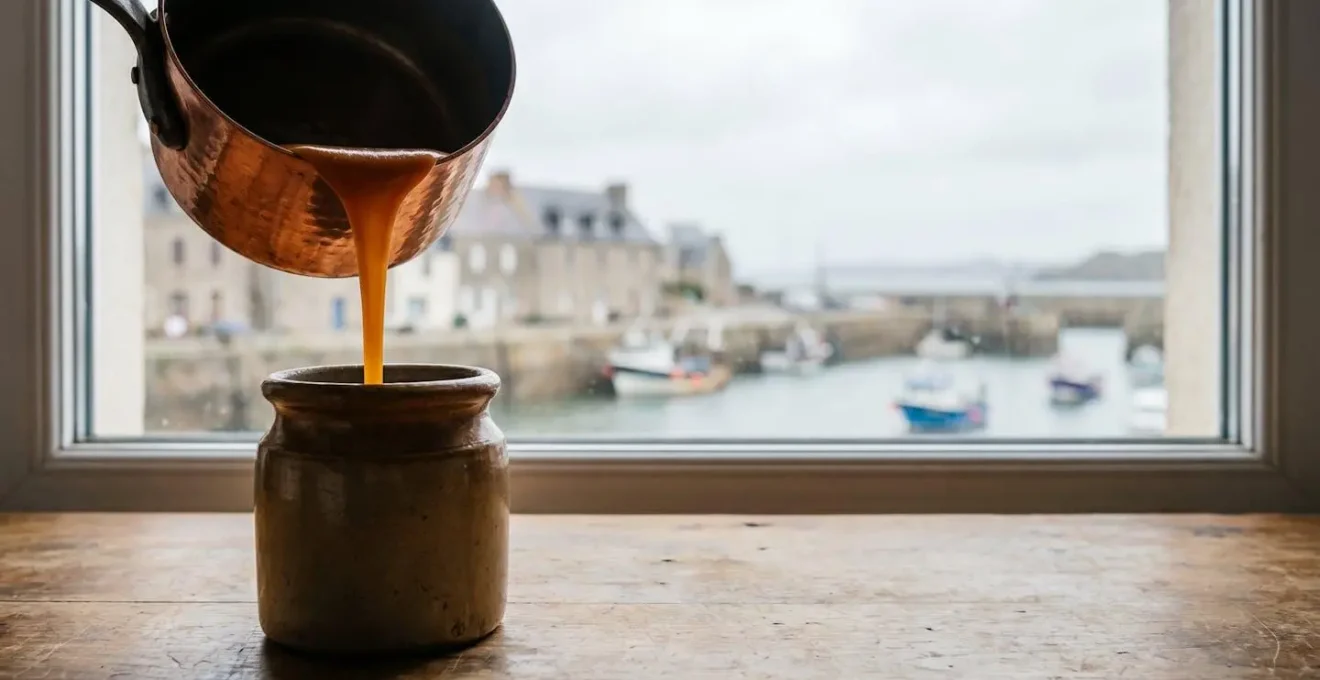 Filet de caramel au beurre salé doré coulant dans un pot en terre cuite, dans une cuisine bretonne baignée de lumière naturelle